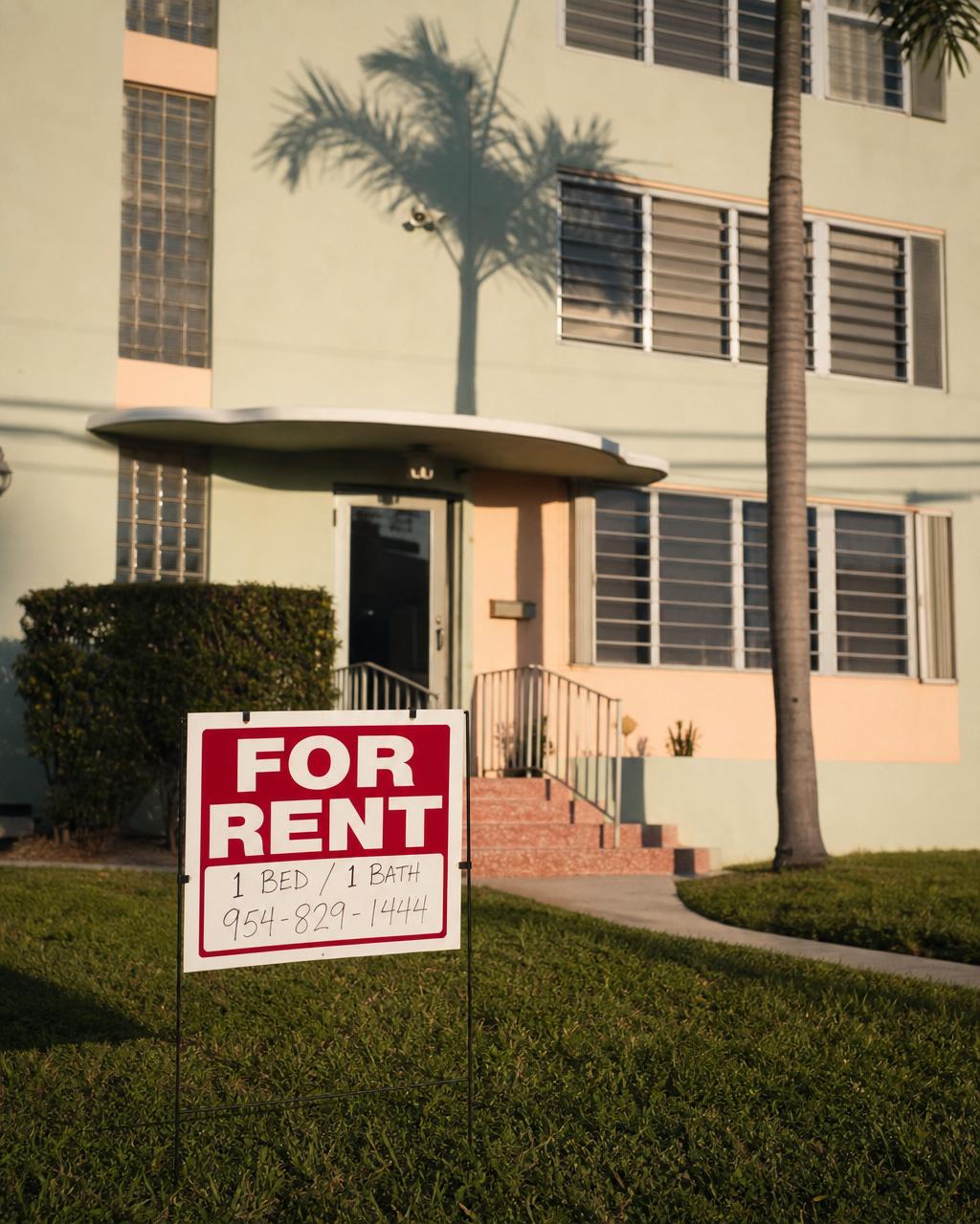 A red-and-white "For Rent" sign in front of a pastel South Florida apartment building